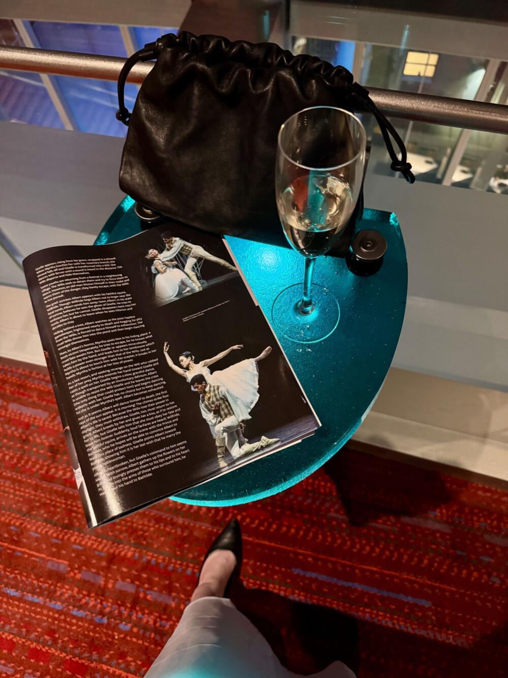 Photo of a Giselle performance program, a flute of champagne, and a black leather clutch bag sitting atop a small blue table in the foyer of a theater. Below the table, one foot wearing a black pump is visible.