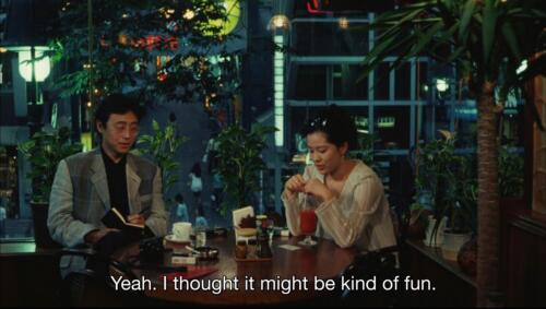 Still frame from the movie A New Love in Tokyo. Two characters, Rei and her interviewer, chat in a quiet kissaten in the neon-lit, Shōwa-vibes, Shibuya evening. The subtitle reads, "Yeah. I thought it might be kind of fun," in reference to her decision to embark on a career as a dominatrix.