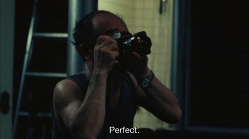Still frame of the photographer Nobuyoshi Araki in the movie A New Love in Tokyo. Araki is kneeling and taking a photo. He wears a tank top. The subtitle reads, "Perfect."