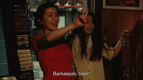 Still frame from the movie A New Love in Tokyo. Rei and Ayumi, two young women, stand in a bar. Rei holds up 2 fingers. The subtitle reads, "Barkeeper, beer!"