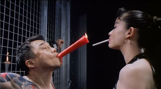 Rei allows her fully restrained submissive client to light her cigarette with a large, red candle held up by his mouth.